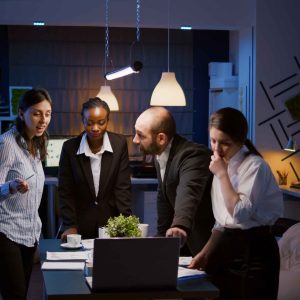 Multi ethnic businesspeople brainstorming company ideas checking graphs statistics using laptop standing at conference table. Diverse teamwork working at management presentation in meeting room