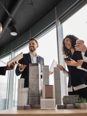 Multiethnic real estate agents, standing near table with 3d city model in office with panoramic city view, while handsome male project manager presenting them report about company's activity on laptop